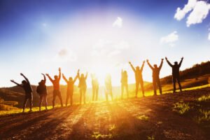Big group of happy friends stands on sunset backdrop with raised arms together. Friendship or teamwork concept.